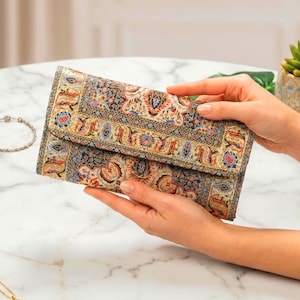 May include: A rectangular wallet featuring a detailed, multi-colored woven pattern reminiscent of a traditional rug. The design incorporates intricate floral and geometric motifs in shades of black, cream, red, blue, and gold, with a flap closure.