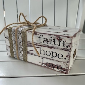May include: A rustic wooden box with a distressed white paint finish. The box is decorated with burlap ribbon, lace, and twine. The words "faith", "hope", and "love" are written on the box in black lettering.