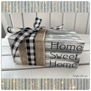 May include: A rustic wooden block with the words "Home Sweet Home" in black lettering. The block is wrapped with a burlap band and a black and white checkered ribbon. The block is painted in shades of gray and white.