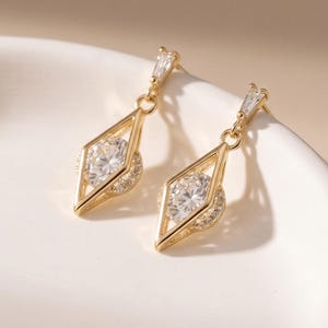 May include: A pair of gold-toned drop earrings featuring a geometric diamond-shaped frame. Each earring contains a central round clear stone surrounded by a small halo of pavé-set crystals, suspended from a thin gold bar with a baguette-cut stone.