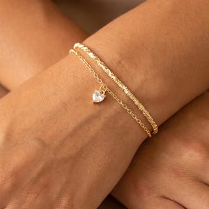 May include: A double-strand gold-toned bracelet featuring one textured chain and one delicate link chain with a small, clear, heart-shaped pendant charm. The jewelry piece is worn around the wrist, showcasing a combination of a twisted metal texture and a smooth, polished finish on the heart-shaped accent.