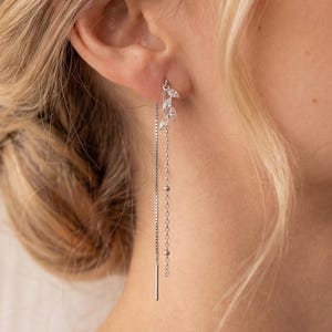 May include: Elegant silver drop earrings with a delicate design. The earrings feature a cluster of small crystals at the ear and a long, dangling chain with small beads and a straight bar at the end. The earrings are worn by a person with blonde hair.