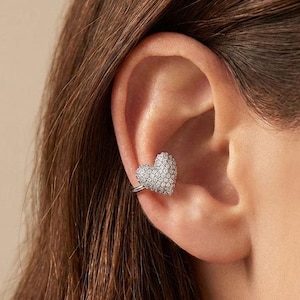 May include: A heart-shaped ear cuff, encrusted with small, sparkling stones, is worn on the ear. The jewelry piece is silver-toned and appears to be made of metal. The ear cuff is a stylish accessory.
