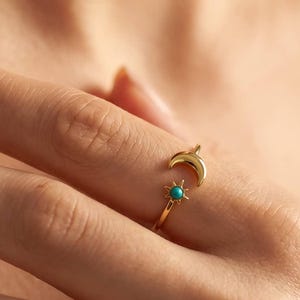 May include: A gold-colored ring featuring a crescent moon and a small turquoise-colored stone. The ring is worn on a finger, with the moon design wrapping around the top of the finger. The ring is a delicate piece of jewelry.
