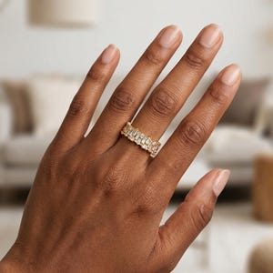 May include: A gold-toned ring with rectangular, clear stones set in a row. The ring is on a person's hand, with the focus on the jewelry. The ring's design is modern and elegant, suitable for various occasions.