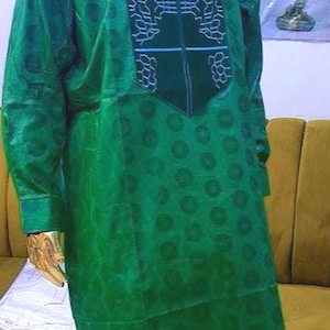 May include: A green, long-sleeved outfit with a decorative pattern on the chest and a matching pair of pants. The outfit is displayed on a gold mannequin. The fabric has a subtle, circular pattern. The outfit is a traditional style.