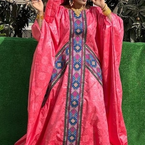 May include: A vibrant pink, flowing dress with a matching head covering. The dress features intricate geometric patterns in blue, black, and pink down the center. Gold jewelry accents the ensemble, creating a striking and elegant look.