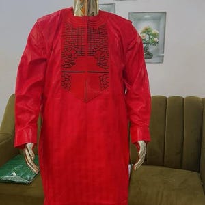 May include: A red, long-sleeved outfit displayed on a gold mannequin. The outfit includes a tunic with embroidered detailing and matching pants. The mannequin is in front of a green sofa and framed wall art.