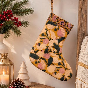 May include: A mustard yellow Christmas stocking featuring a repeating botanical print of dark green leaves, light pink circular fruits, and small berries. The stocking has a matching fabric loop at the top for hanging and a wide cuff at the opening.