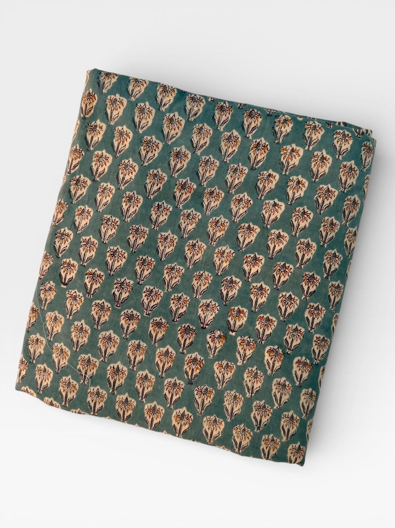 A neatly folded rectangular piece of teal blue fabric with a repeating pattern of light brown and white floral motifs is centered on a white background.