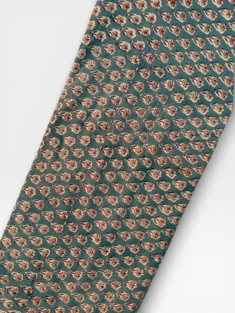 A long, narrow strip of teal blue fabric with a repeating pattern of light brown and white floral motifs is laid diagonally across a white background.