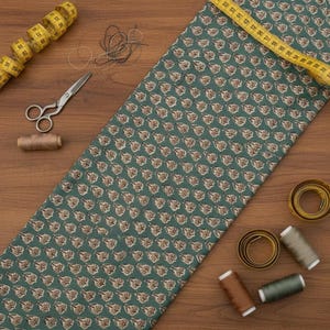 A teal blue cotton fabric with light brown and white floral motifs, laid on a wooden surface with sewing notions including scissors, thread, and a measuring tape.