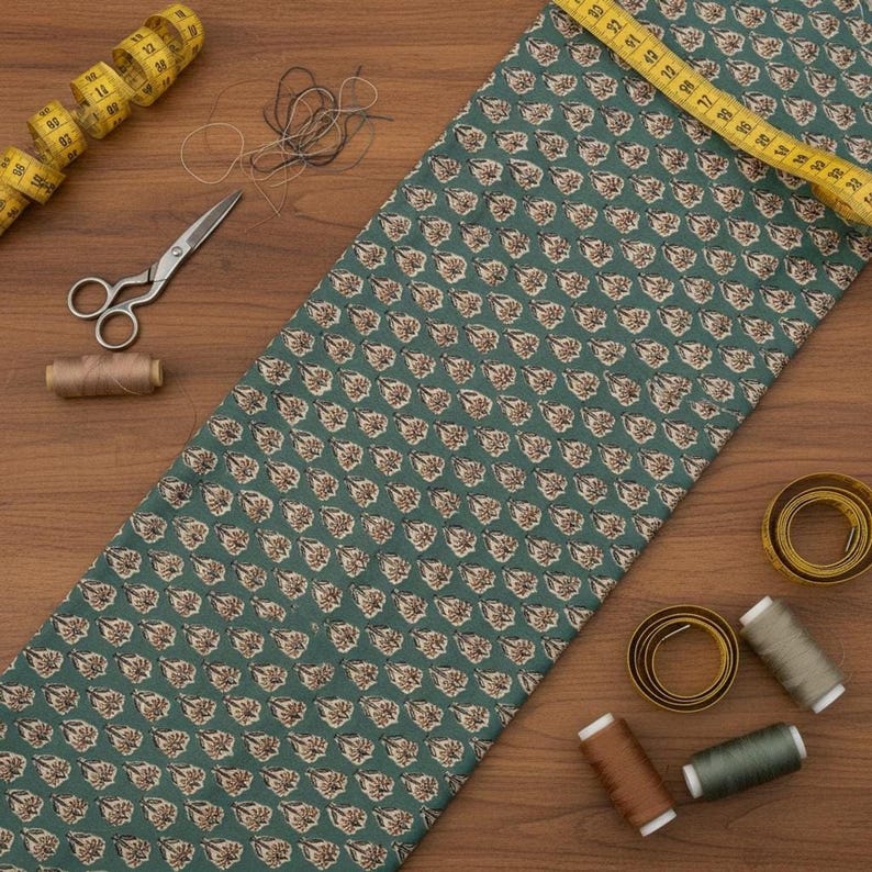 A teal blue cotton fabric with light brown and white floral motifs, laid on a wooden surface with sewing notions including scissors, thread, and a measuring tape.
