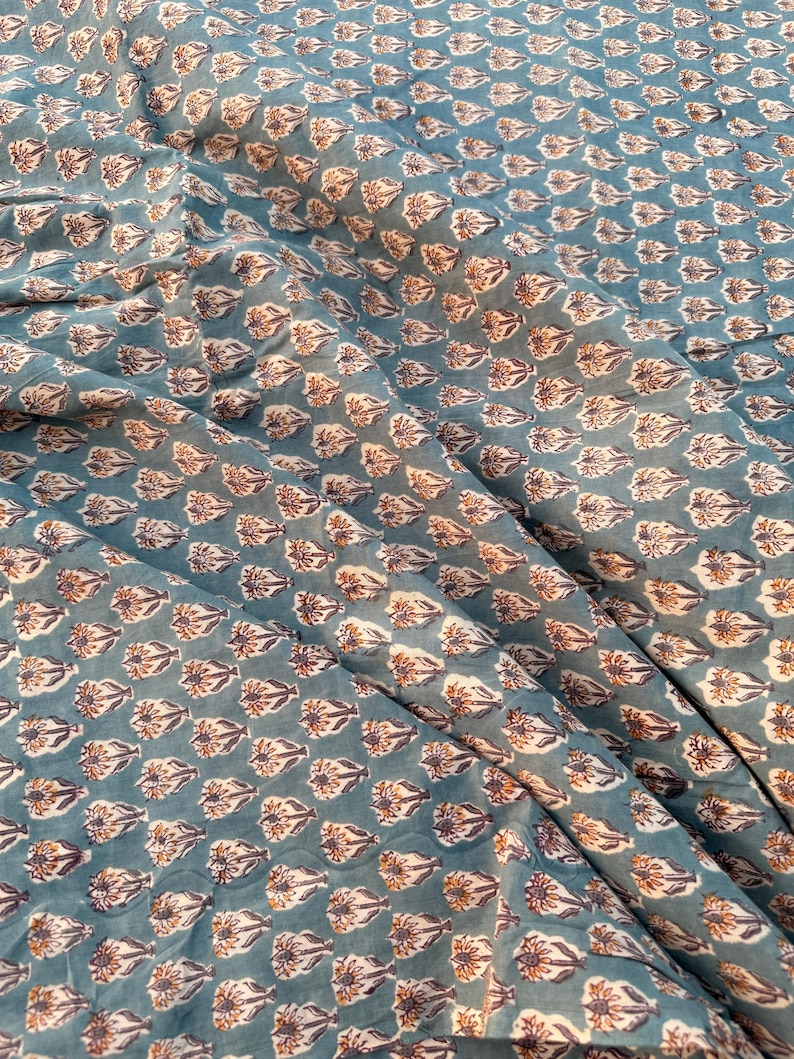 A teal blue cotton fabric with light brown and white floral motifs, draped with soft folds and shadows.