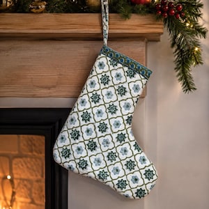 May include: A quilted Christmas stocking featuring a white base with a repeating pattern of dark green and light blue floral motifs enclosed in thin green grid lines. The stocking has a matching patterned cuff and a fabric loop for hanging.