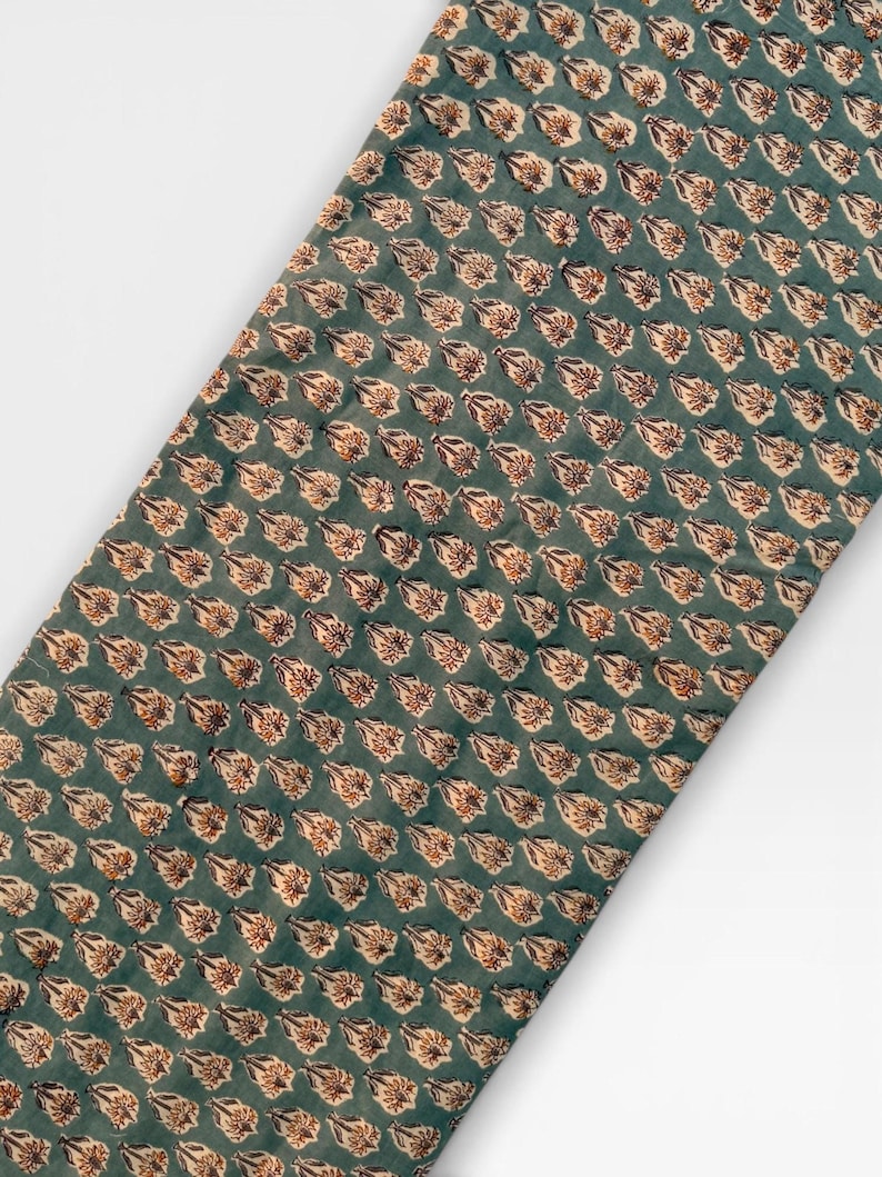 A long, narrow strip of teal blue fabric with a repeating pattern of light brown and white floral motifs is laid diagonally across a white background.