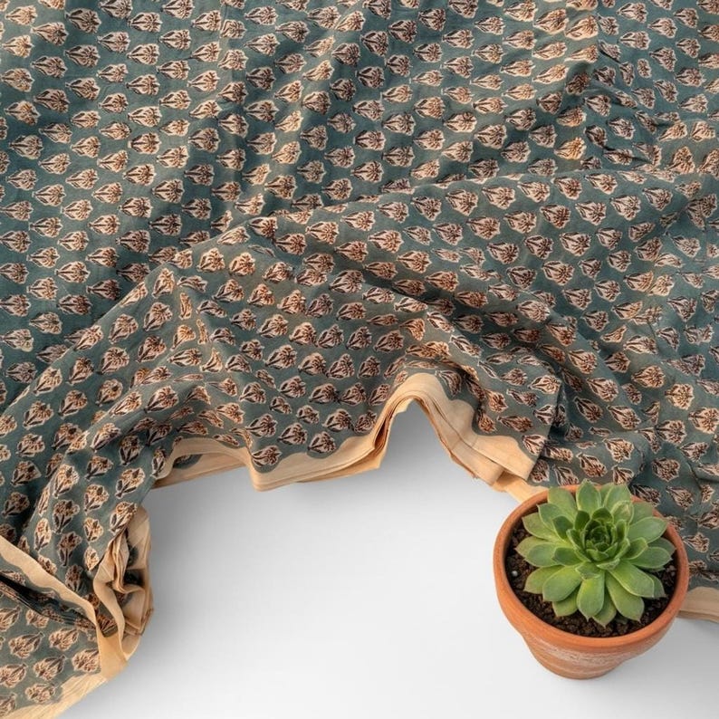 A teal blue fabric with a repeating pattern of small, light brown and white floral motifs is draped next to a small green succulent in a terracotta pot.