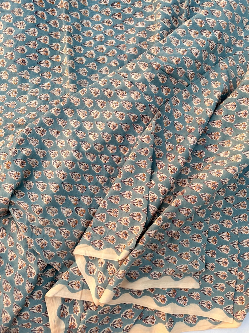 A teal blue cotton fabric with light brown and white floral motifs, draped with several soft folds and a visible white edge.