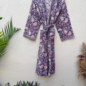 Puede incluir: Bata con estampado floral púrpura con cinturón a juego, colgada de una percha de madera. La bata presenta un patrón repetido de flores rosas y blancas sobre un fondo púrpura oscuro. Fabricada en tejido suave y ligero.