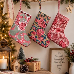 Puede incluir: Tres calcetines navideños de tela colgados de una rama de abedul. El calcetín izquierdo es rosa con motivos florales blancos y verdes. El central es azul verdoso con diseños florales rosas y dorados. El derecho es rojo con motivos florales rosas.