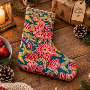 Op de afbeelding: Een kerstkous van stof met een blauwgroene achtergrond en een herhalend patroon van grote roze bloemen en groene bladeren. De kous heeft een bijpassende stoffen lus aan de bovenkant om op te hangen en een decoratieve rand langs de boord.