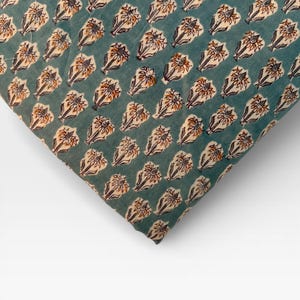 A corner of a neatly folded teal blue fabric with a repeating pattern of light brown and white floral motifs is shown on a white background.