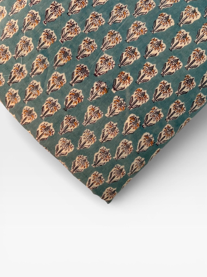 A corner of a neatly folded teal blue fabric with a repeating pattern of light brown and white floral motifs is shown on a white background.