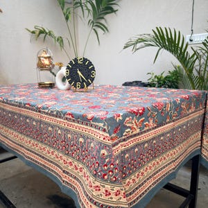 Könnte beinhalten: Ein rechteckiges Tischtuch mit floralem Muster in Blau, Rot und Creme. Auf dem Tisch befindet sich eine schwarze Uhr mit goldenen Zeigern und einem goldenen Ständer. Eine weiße Tasse und eine kleine goldene Teekanne sind ebenfalls zu sehen. Das Tischtuch ist ca. 150 cm x 200 cm groß.