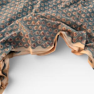 A teal blue cotton fabric with light brown and white floral motifs, folded and draped over a white surface, showing a light tan underside.