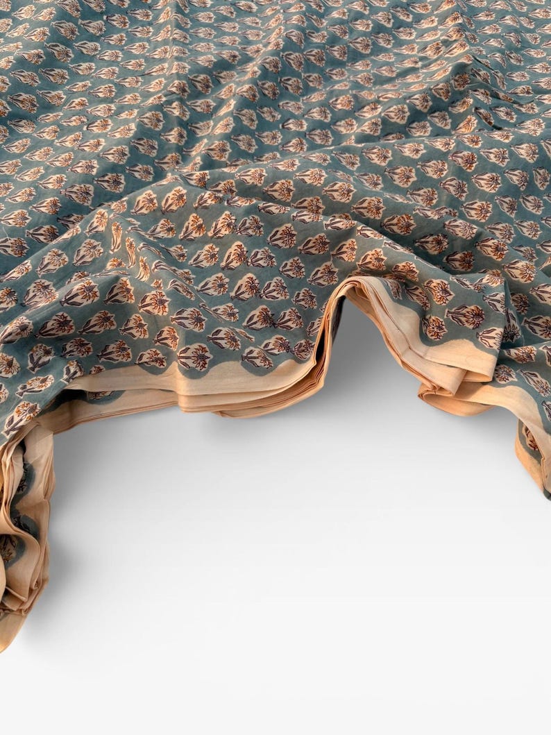 A teal blue cotton fabric with light brown and white floral motifs, folded and draped over a white surface, showing a light tan underside.