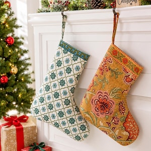 May include: Two fabric Christmas stockings hanging from a white mantel. The left stocking features a white background with a blue and green geometric floral pattern. The right stocking has a mustard yellow background with a multi-coloured floral print.