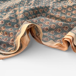 A teal blue cotton fabric with light brown and white floral motifs, folded and draped in a wave-like pattern over a white surface, showing a light tan underside.