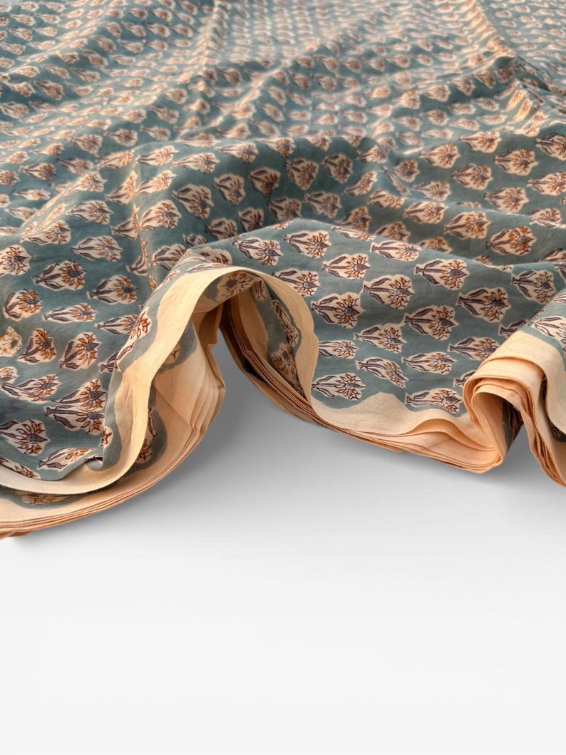 A teal blue cotton fabric with light brown and white floral motifs, folded and draped in a wave-like pattern over a white surface, showing a light tan underside.