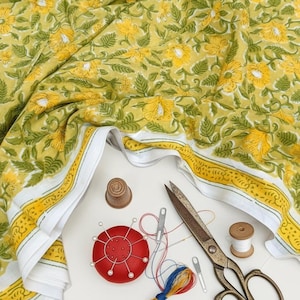 May include: Yellow floral fabric with green leaves and white borders, with sewing tools: scissors, a red pin cushion, thimble, needles, and thread spools. The fabric is suitable for crafting or home decor.