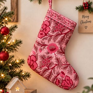 Puede incluir: Un calcetín navideño de tela rosa con un patrón floral repetitivo en tonos rosa oscuro y burdeos. El calcetín tiene un lazo de tela a juego en la parte superior para colgarlo y un detalle de borde decorativo a lo largo del puño superior.