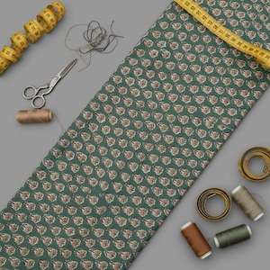 A teal blue fabric with light brown and white floral motifs is laid flat with a yellow measuring tape, scissors, and spools of thread on a gray background.