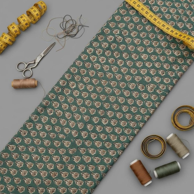 A teal blue fabric with light brown and white floral motifs is laid flat with a yellow measuring tape, scissors, and spools of thread on a gray background.