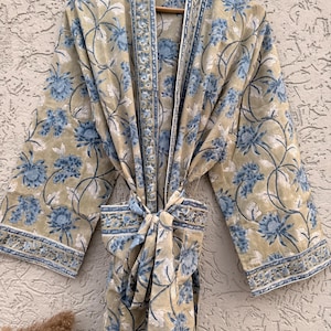 May include: A beige floral patterned robe with blue and white accents. The robe is hanging on a wooden hanger and tied with a matching belt. The floral pattern is repeated throughout the robe's design.