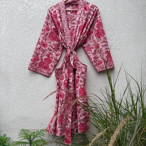 May include: A long-sleeved robe featuring a light pink base with a repeating dark pink floral pattern. The garment includes a shawl collar, wide sleeves, and a matching fabric tie belt at the waist, all hanging from a wooden hanger.