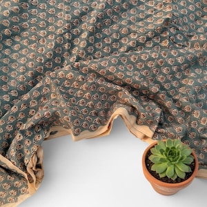 May include: A teal fabric with a repeating pattern of brown and white floral shapes. The fabric has a cream-colored border and is draped over a white surface. A small potted succulent plant sits in the lower right corner.