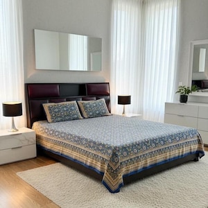 May include: A bedroom with a bed dressed in a blue and gold patterned bedspread and pillowcases. The bed has a dark headboard, white bedside tables with lamps, and a white rug. Sheer curtains frame the scene. The room is decorated in neutral tones.