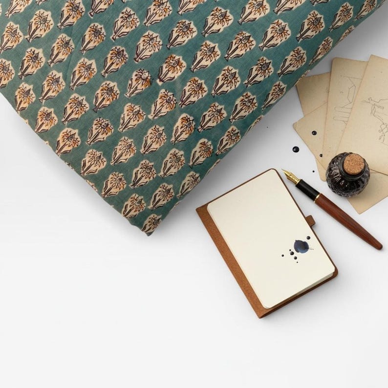 A neatly folded stack of teal blue fabric with light brown and white floral motifs is next to a small notebook, an inkwell, and a pen on a white surface.