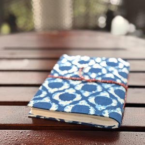 Puede incluir: Un pequeño diario cerrado con una cubierta estampada en azul y blanco. El diseño de la cubierta presenta un patrón repetitivo de círculos conectados. Una cuerda roja envuelve el diario para mantenerlo cerrado. El diario descansa sobre una superficie de madera oscura.