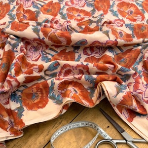 May include: Cream-colored fabric with a repeating pattern of orange and pink poppies and teal leaves. A measuring tape and scissors are on a wooden surface, suggesting a sewing or crafting project.