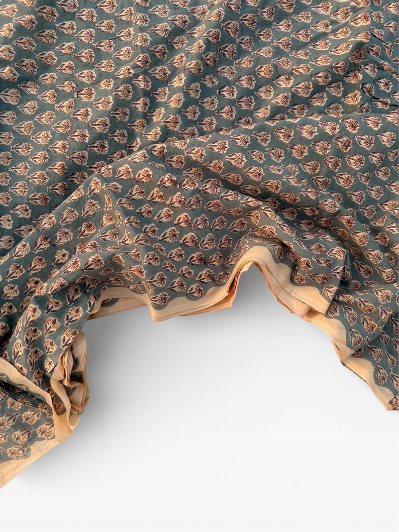 A teal blue cotton fabric with light brown and white floral motifs, draped over a white surface, revealing a light tan underside.