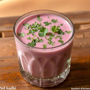 May include: A clear glass tumbler filled with a smooth, opaque pink liquid, topped with fresh green cilantro leaves. The glass features a decorative scalloped base design. The text Sol Kadhi and Sunita's Kitchen is visible at the bottom.