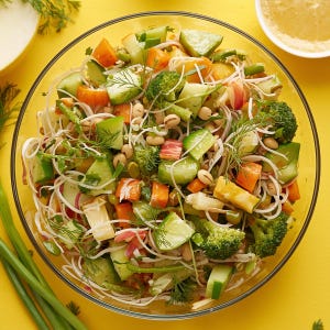 May include: A colorful salad in a clear glass bowl. The salad contains cucumbers, carrots, broccoli, beans, and noodles. Fresh dill is sprinkled throughout. The bowl sits on a yellow surface, with a bowl of dressing and green onions in the background.