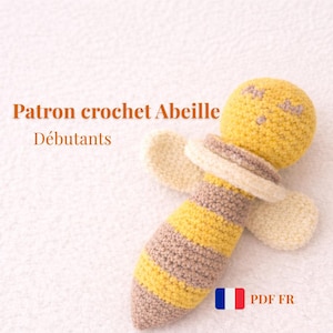 Peut inclure: Un jouet d'abeille crocheté à la main, jaune, beige et crème. L'abeille a une tête ronde, un corps rayé et deux ailes crème. Le texte "Patron crochet Abeille Débutants" est visible.