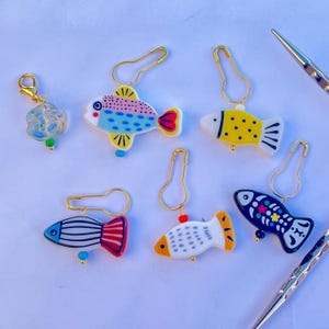 May include: A collection of colorful fish-shaped stitch markers with gold-tone safety pin attachments. The fish feature various designs, including polka dots, stripes, and floral patterns. A clear, rose-shaped charm is also included.