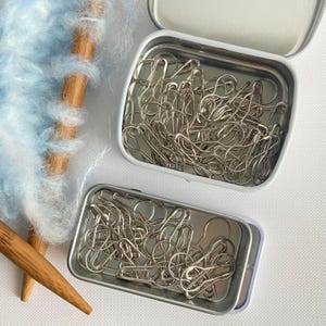 May include: Two small, open, rectangular metal tins filled with silver-colored safety pins. A bundle of light blue yarn and two wooden knitting needles are in the background. The tins are on a white surface.